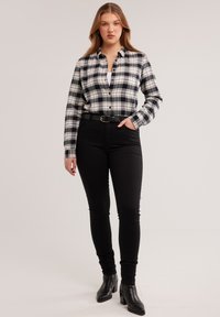 Noor naine, kes kannab must-valget ruudulist särki, musti kõrge vöökohaga pükse, musta vööd ja musti pahkluu saabastega, seisab käega taskus.