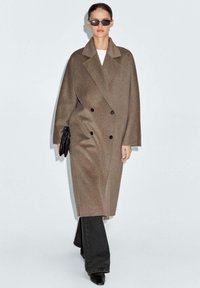 Cappotto di lana marrone doppiopetto con ampi rever, spacco laterale e bottoni neri, abbinato a jeans neri a zampa d'elefante e occhiali da sole.