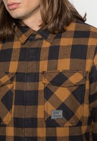 Flanellskjorta med ett svart och orange rutmönster, två bröstfickor och en märkesetikett på den vänstra fickan. Tillverkad av strävt tyg.