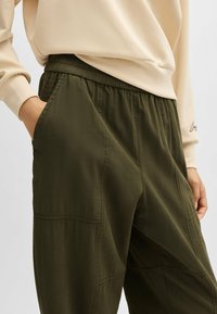 Olijfgroene, lichtgewicht broek met elastische tailleband, zijzakken, gestructureerde stof en subtiele stikdetails. Gedragen met een beige lange mouwen top.