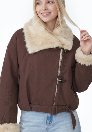 Donna sorridente che indossa una giacca marrone con colletto e polsini in pelliccia beige, che tiene una ciocca dei suoi capelli biondi, indossa jeans azzurri.