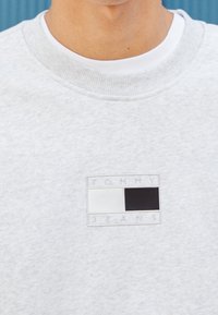 Sudadera gris claro con una tela texturizada. Presenta un logotipo de marca rectangular en blanco y negro en el pecho, con cuello redondeado.