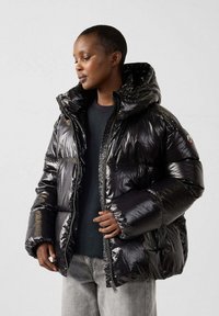 Chaqueta acolchada negra y brillante con capucha, con cremallera frontal, grandes bolsillos y un diseño acolchado. Lleva puesta sobre una camiseta oscura y unos vaqueros.