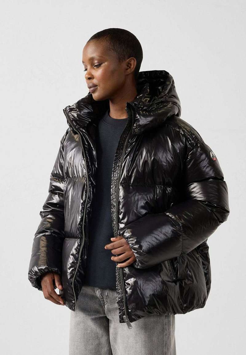 Chaqueta acolchada negra y brillante con capucha, con cremallera frontal, grandes bolsillos y un diseño acolchado. Lleva puesta sobre una camiseta oscura y unos vaqueros.