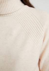 Kremowy prążkowany sweter z golfem na osobie, ukazujący szczegółowy wzór dzianiny na ramieniu i w okolicy kołnierza.