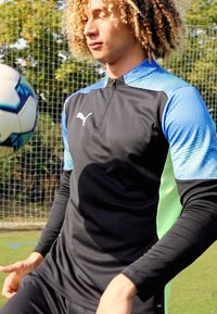 Schwarzes Sportshirt mit blauen Akzenten und einem mintgrünen Seiteneinsatz. Das Shirt hat einen Reißverschlusskragen und ein strukturiertes Material. Ein Fußball ist in Bewegung.