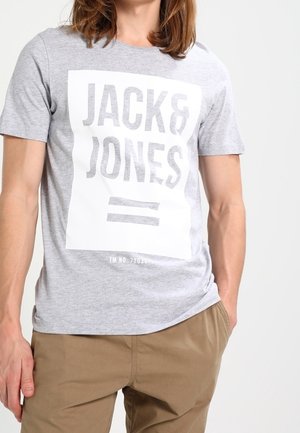 Mand iført en lysegrå T-shirt med "JACK & JONES" trykt med store bogstaver inden for en hvid rektangel, kombineret med khakifarvede bukser.