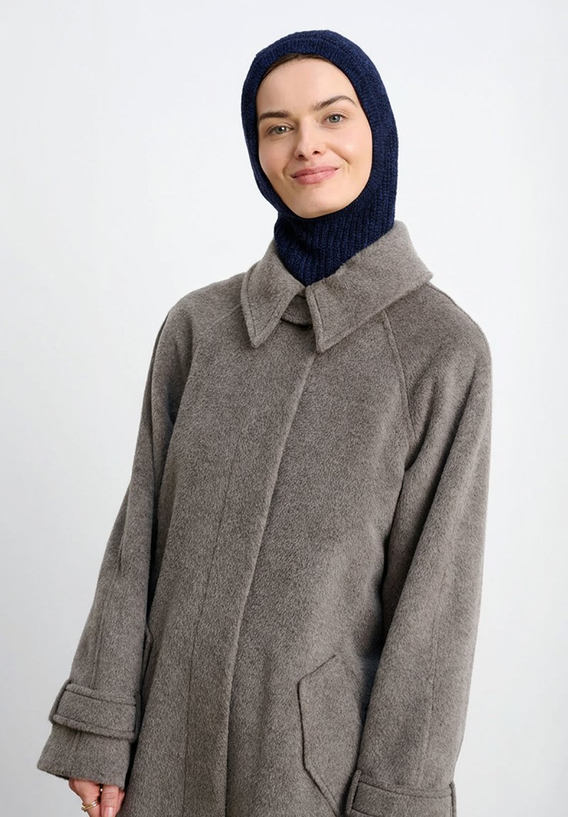 Szary wełniany płaszcz z kołnierzem i bocznymi kieszeniami, zestawiony z granatowym prążkowanym dzianinowym szalikiem na głowę, charakteryzujący się luźnym krojem i miękką fakturą.