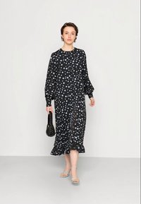 Vestido preto de mangas longas com decote redondo, apresentando um padrão de bolinhas brancas e azul claro e uma bainha com babados. Usado com sandálias de tiras.