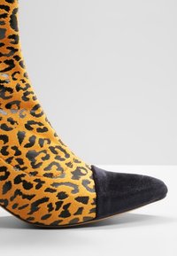 La chaussure à talons aiguilles en imprimé léopard présente un dessus en daim orange avec des taches noires et un capuchon de pointe en daim noir. Texture lisse dans l'ensemble.