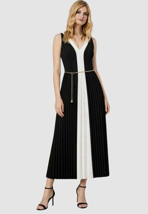 Donna che indossa un abito lungo plissettato senza maniche, bianco e nero, con scollatura a V e una sottile cintura a catena dorata, abbinato a sandali neri con tacco alto.