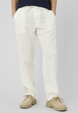 Personne portant un pantalon en lin blanc à fines rayures avec taille à cordon et des chaussures beiges à semelle épaisse et lacées, mains dans les poches.