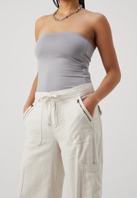 Šedý top bez ramínek s hladkou texturou, v kombinaci s bílými cargo kalhotami na stahovací šňůrku, které mají zipové kapsy a volný střih.