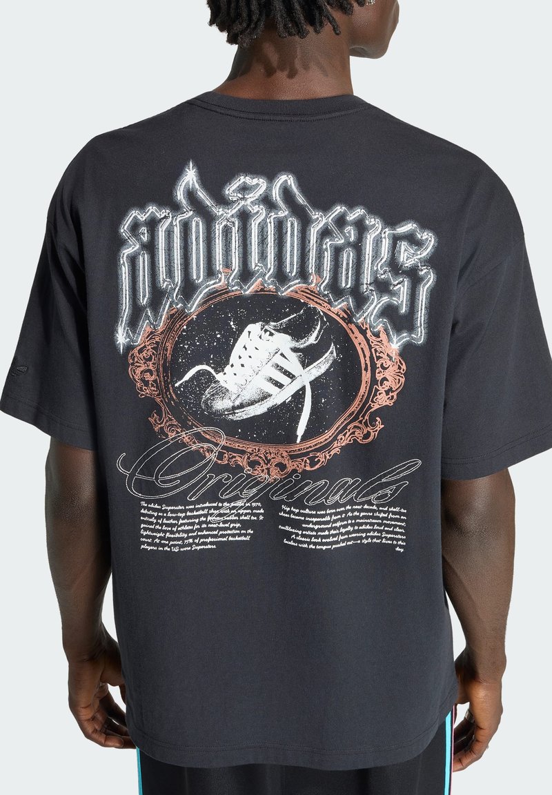 Persona que lleva una camiseta negra con un gran gráfico de una zapatilla blanca enmarcada por un diseño adornado y el texto "adidas Originals" en la espalda.