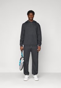 Uomo che indossa un felpa grigia scura e pantaloni della tuta, con in mano una racchetta da tennis nera e blu, in piedi contro uno sfondo chiaro e semplice.