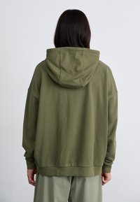 Ελαφρώς υπερμεγέθη φόρμα σε ελιά με κορδόνι στην κουκούλα, ριμπ μανσέτες και στρίφωμα. Λείο ύφασμα από βαμβακερό μίγμα, μινιμαλιστικός σχεδιασμός και άνετη εφαρμογή.