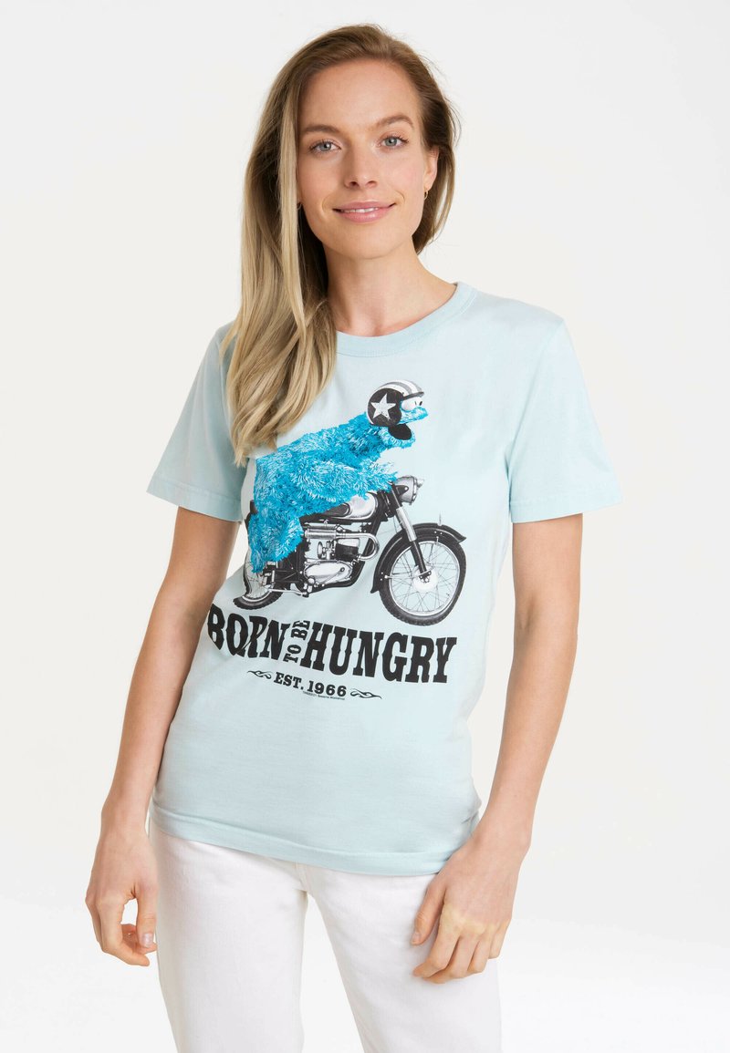 Hellblaues T-Shirt mit einem bunten Grafikmotiv eines pelzigen Charakters auf einem Motorrad, mit schwarzem Text "BORN TO BE HUNGRY".