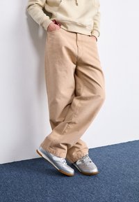 Pantalones beige de corte holgado con textura suave, combinados con zapatillas deportivas plateadas y una sudadera con capucha de color beige claro. La escena presenta una alfombra azul.
