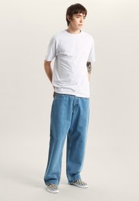 Ljusgrå kortärmad t-shirt med avslappnad passform, kombinerad med löst sittande blå denimjeans. Svartvita rutiga slip-on-skor. Minimalistisk design.