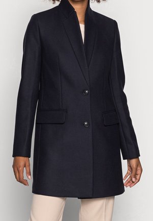 Femme portant un manteau noir sur mesure avec col à revers, deux boutons et poches avant à rabat, associé à un pantalon clair.
