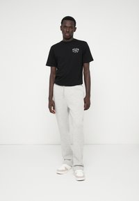 Μαύρο κοντομάνικο μπλουζάκι με λογότυπο, συνδυασμένο με ανοιχτό γκρι φόρμες. Μοντέλο που φοράει λευκά και μπεζ αθλητικά παπούτσια, στέκεται μπροστά σε ουδέτερο φόντο.