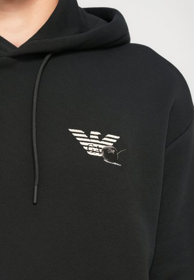 Melna kapuce ar baltu svītru ērgļa logotipu un tumši ķiršu izšuvumu uz krūtīm.