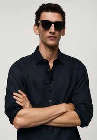 Camicia nera con maniche arrotolate, abbinata a occhiali da sole neri. Il modello ha le braccia incrociate e mostra un'espressione neutra.