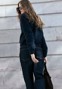 Ciemnoniebieski sweter o teksturowanym wykończeniu w połączeniu z wysokim stanem dżinsów w ciemnym kolorze. Model trzyma brązową torbę na ramię, odwracając się plecami.