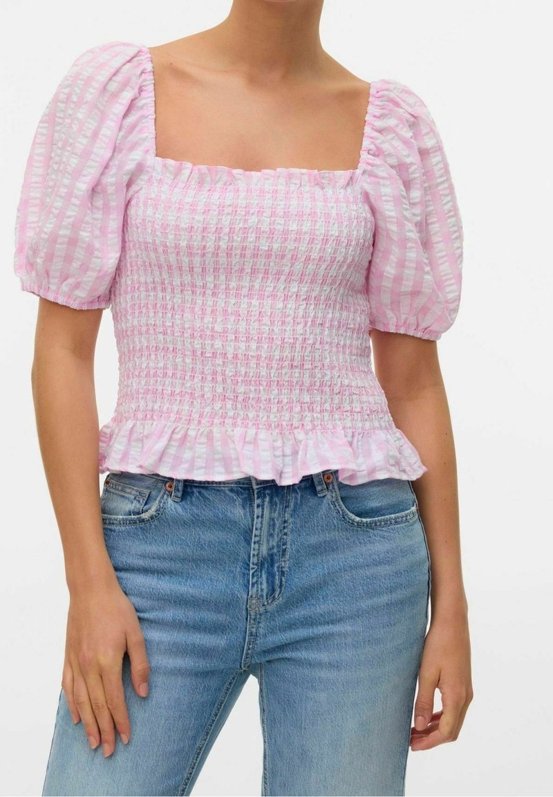 Femme portant un haut froncé à rayures roses et blanches avec manches bouffantes et ourlet à volants, associé à un jean en denim bleu.