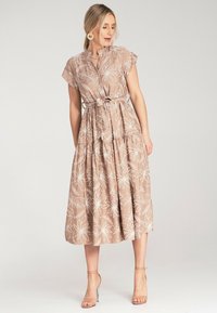 Hellbraunes Maxikleid mit weißen Blumenstickereien, kurzen Ärmeln, einem Tunikabund und einem gestuften Rock. Kombiniert mit klaren, hochhackigen Sandalen.