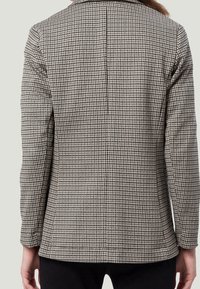 Marynarka w czarno-białą kratę houndstooth z klapami, prostym krojem i poduszkami na ramionach. Widok z tyłu ukazuje czyste linie i szwy.