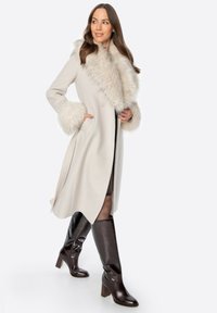 Beżowy wełniany płaszcz z futrzanym kołnierzem i mankietami, o dopasowanym kroju i zapinany na jeden guzik. Brązowe skórzane buty za kolano.