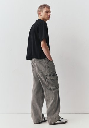 Fiatal férfi rövid hajjal, fekete túlméretezett pólót, széles szürke cargo nadrágot és fehér tornacipőt visel, sima fehér fal előtt áll.