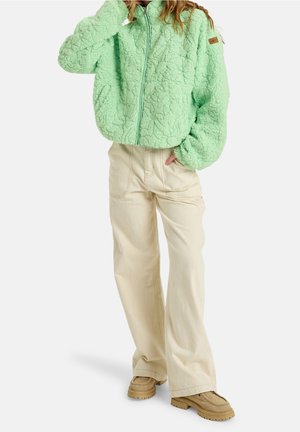 Osoba ubrana w miętowo-zieloną teksturowaną kurtkę na zamek, beżowe szerokie spodnie i brązowe platformy, stojąca z jedną ręką w kieszeni.