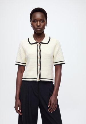 Donna con capelli corti che indossa un cardigan bianco a manica corta in maglia con rifiniture nere e pantaloni neri a gamba larga, in piedi su sfondo bianco.