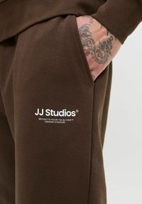 Smeđe pamučne jogger hlače s bočnom džepom, ukrašene bijelim otisnutim logotipom "JJ Studios" i suptilnim tekstualnim detaljima. Glatka tekstura tkanine.