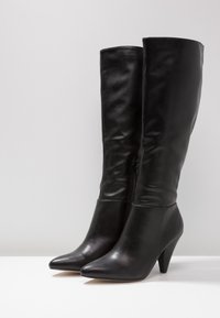 Ein Paar schwarze knielange Lederstiefel mit spitzen Zehen und mittelhohen Kegelhufen auf einer weißen Oberfläche.