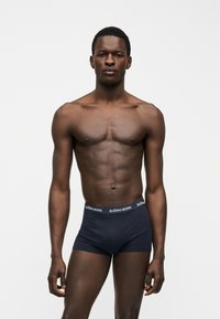 Αθλητικός άντρας όρθιος με το σώμα μπροστά, φοράει σκούρα ναυτικά μπόξερ με τη ζώνη μέσης διακοσμημένη με το λογότυπο "BJÖRN BORG", σε απλό λευκό φόντο.