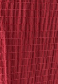 Texturovaná tkanina v tmavě červené barvě s mřížkovým vzorem, zdůrazňující měkké, tkané vlákna a bohatý odstín, který naznačuje teplo a pohodlí.
