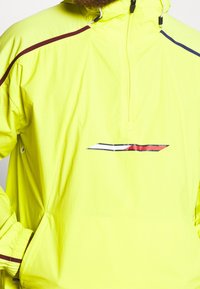 Lys gul vindjakke med halv-zip design, med maroon og marineblå detaljer langs skuldrene og et branding grafisk element.