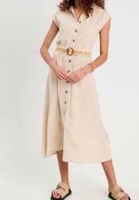 Robe chemise en lin beige à manches courtes avec deux poches avant, cintrée par une ceinture à motif. Modèles assortis avec des sandales ornées.