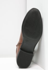Chaussure en cuir marron avec une semelle en caoutchouc noir texturé. Présente un détail tressé sur le côté et un design à bout rond.