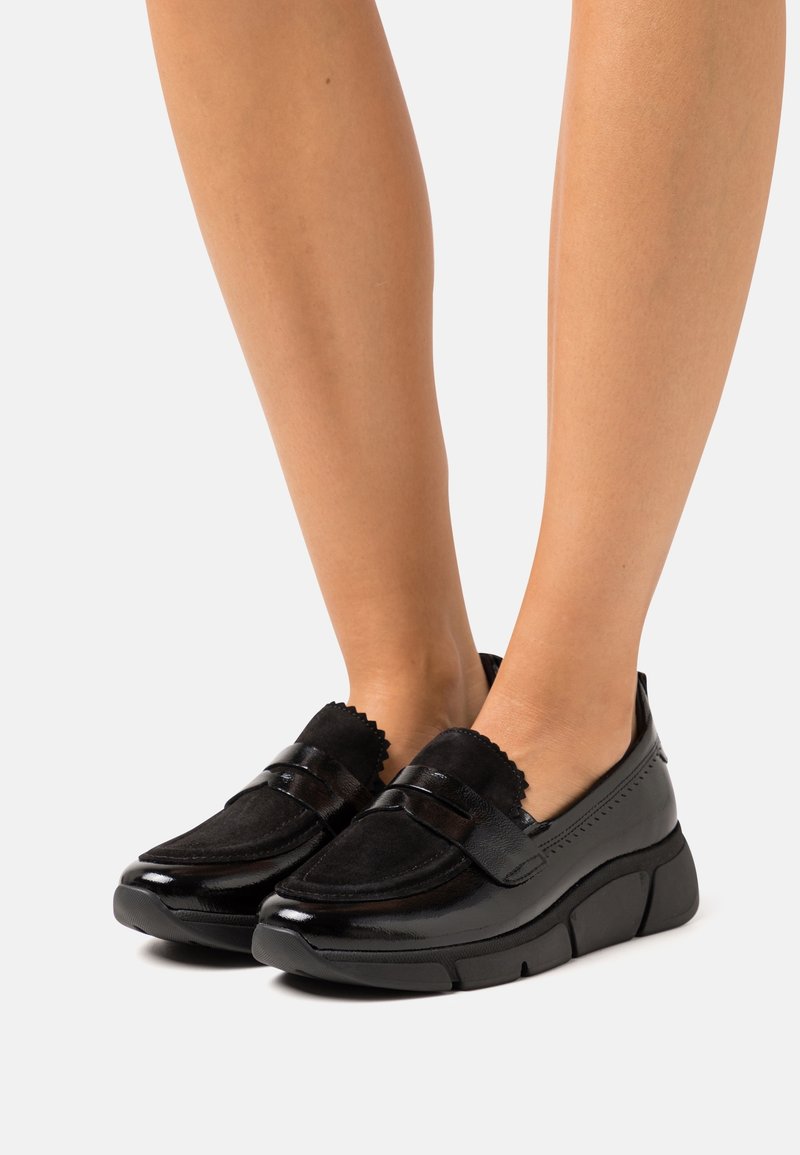 Mocassins en cuir noir et daim avec semelles en caoutchouc épaisses portés sur pieds nus sur fond blanc.