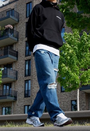 Osoba ubrana w czarną bluzę z kapturem, białe trampki i podarte niebieskie dżinsy spacerująca na zewnątrz w pobliżu nowoczesnego budynku mieszkalnego i zielonego drzewa.