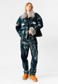 Chaqueta de denim con cuello de piel sintética gris, patrones abstractos en azul y verde, combinada con pantalones de chándal a juego y zapatos de suela gruesa en color naranja.