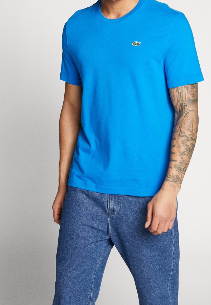 Homme portant un t-shirt Lacoste bleu vif et un jean bleu, avec une manche de tatouage détaillée visible sur son bras droit.