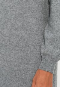 Mâna persoanei se sprijină pe un pulover tricotat moale, gri, cu mâneci lungi și mansete cu elastic, evidențiind textura țesăturii și modul în care se potrivește articolul vestimentar.