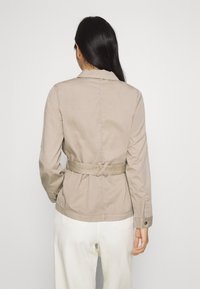 Beige jakke med bælte, lange ærmer, krave og tekstureret stof. Bagfra ses en indsnævret talje og knaplukninger på ærmekanterne.