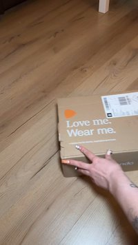 Brown cardboard box with white lettering saying "Love me. Wear me." and an orange triangle logo. A hand is reaching for the box.