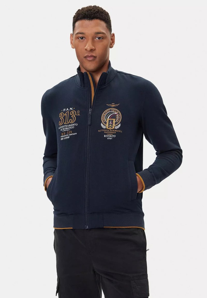 Felpa zip con cappuccio navy con grafiche ricamate, colletto interno arancione a contrasto e tasche frontali. Tessuto morbido e design aderente.